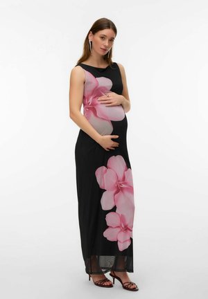 Femme enceinte portant une robe noire sans manches à grands motifs floraux roses, portant des boucles d'oreilles et des sandales à talons, les mains caressant son ventre.