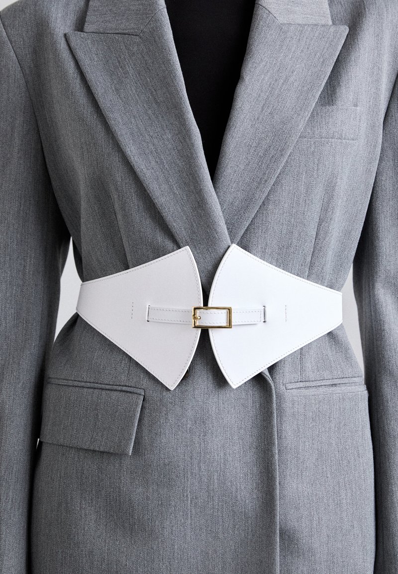 Cinturón de cuero blanco con hebilla dorada, con un diseño ancho y puntiagudo, llevado sobre un blazer gris de tela texturizada y con detalle de bolsillo.