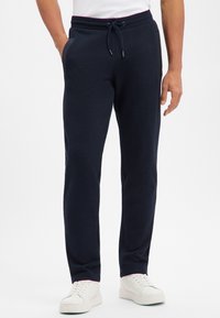 Mężczyzna stojący w czarnych, casualowych spodniach z sznurkiem i białych trampeczkach, ręce włożone do kieszeni, na jednolitym białym tle.