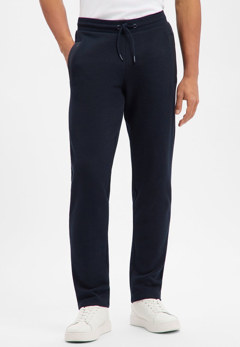 Mężczyzna stojący w czarnych, casualowych spodniach z sznurkiem i białych trampeczkach, ręce włożone do kieszeni, na jednolitym białym tle.