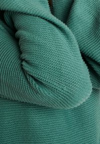 Turkos stickad tyg med en texturerad mönster, med en vikt design och lätta rynkor vid sömmarna, vilket ger en mysig känsla.