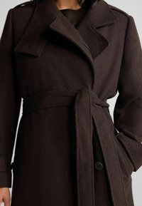 Trench coat di lana marrone con vita cintrata, ampi risvolti e finitura testurizzata. Presenta un bottone nero singolo e tasche laterali.