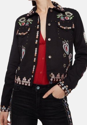 Schwarze Jacke mit Knöpfen, verziert mit bunten Pailletten und bestickten Augen, Blumen und Sternen, getragen über einem roten Top mit schwarzen Hosen.