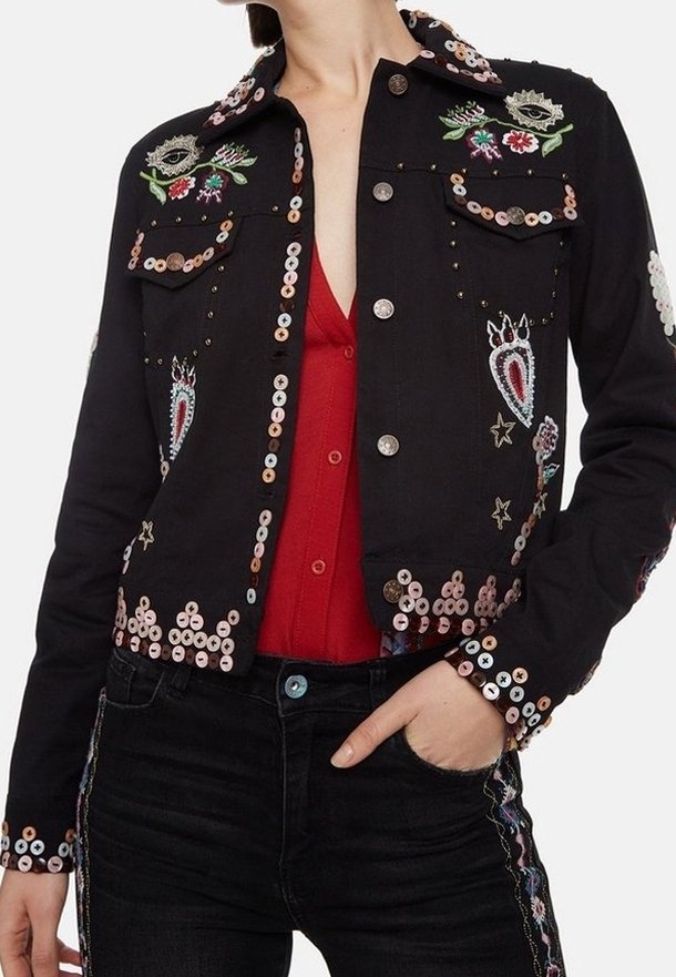 Veste noire boutonnée décorée de sequins colorés et d'yeux, fleurs et étoiles brodés, portée sur un top rouge avec un pantalon noir.