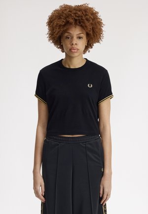 Femme aux cheveux bouclés portant un t-shirt court noir avec un bord doré et un pantalon noir assorti avec des détails dorés, debout devant un fond blanc.