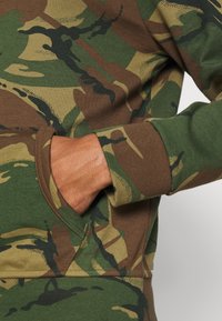 Sudadera de camuflaje verde y marrón con un tejido texturizado, puños acanalados y detalle de bolsillo en el lateral.