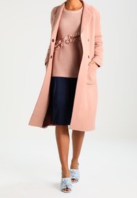 Jasnoróżowy wełniany płaszcz o kroju dwurzędowym, noszony na fakturowanym różowym swetrze i granatowej plisowanej spódnicy. Jasnoniebieskie obcasy zawiązywane na supeł.