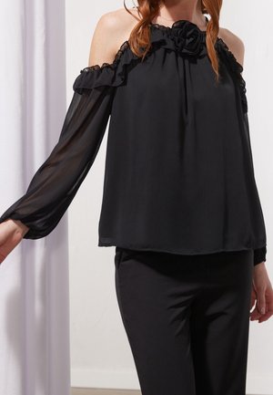 Femme portant un chemisier noir épaules dénudées avec manches longues transparentes et encolure à volants, associée à un pantalon noir, debout devant un fond uni.