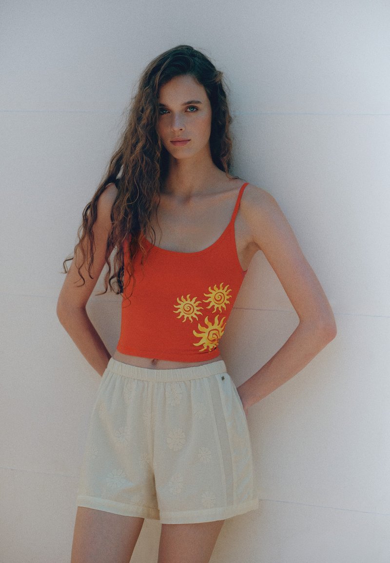 Jeune femme aux longs cheveux bouclés portant un crop top rouge avec des soleils jaunes et un short couleur crème, debout contre un mur clair uni.