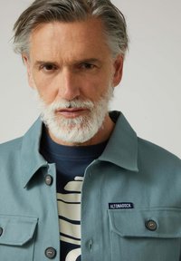 Chaqueta de color azul verdoso claro con tela texturizada, cuello abotonado y bolsillos. Se lleva sobre una camisa azul oscuro con un patrón crema.