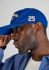 Caciulă de baseball albastră cu broderie roșie și albă și numărul "25" pe lateral. Are un design structurat și o margine curbă.