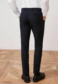 Mørkeblå skræddersyede bukser med slim fit, to baglommer med knapper og en glat tekstur, parret med sorte snørebåndssko.