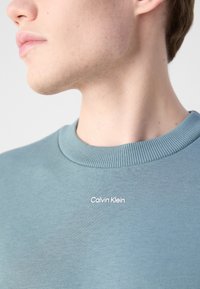 Sudadera azul claro con cuello redondo acanalado. Presenta un logo blanco de "Calvin Klein" en el frente, mostrando una textura de tela suave.