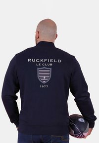 Homme portant une veste sombre avec "Ruckfield Le Club 1977" et un logo en forme de blason dans le dos, tenant un ballon de rugby à son côté.