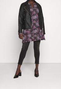 Nainen, joka on pukeutunut mustaan nahkatakkiin, musta-pinkki pilkulliseen tunikaan, mustiin lyhyisiin skinny-farkkuihin ja mustiin kärkinumeroihin, seisoo valkoista taustaa vasten.