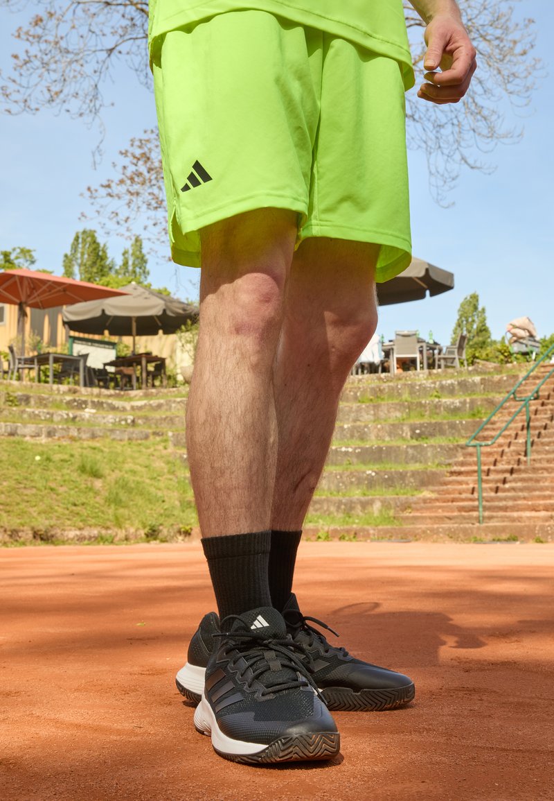 Pantalones cortos deportivos verde neón con acentos negros, combinados con zapatillas negras con suelas blancas y tres rayas negras, llevados con calcetines negros.
