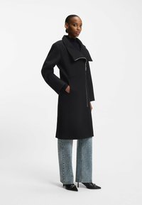 Zwarte jas met een grote kraag en een rits aan de voorkant, met zijzakken. Gecombineerd met lichtblauwe flared jeans versierd met kleine studs.