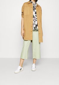 Chemise oversize couleur cuir tanné, blouse blanche à fleurs, pantalon court vert menthe et baskets blanches avec accents noirs. Textures lisses et design décontracté.