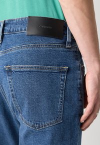 Sötétkék farmernadrág fekete bőr Calvin Klein logórésszel a derekán, hátsó zsebekkel és jól látható varrási részletekkel.
