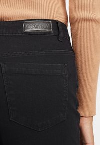 Fekete farmernadrág jellegzetes formával, bőr címkével, amelyen az "OXMO" felirat szerepel, és kontrasztos varrással a hátsó zsebén.