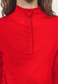 Top rosso a maglia con collo alto e sei bottoni abbinati sulla parte anteriore. Il tessuto appare morbido e liscio con un colore uniforme.