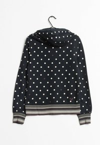 Sudadera azul marino con lunares blancos, puños y cintura a rayas, colgando en una percha negra sobre un fondo blanco.