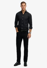 Zwarte button-up overhemd met lange mouwen, gecombineerd met zwarte jeans. Het model draagt een zwarte riem en glanzende zwarte nette schoenen.