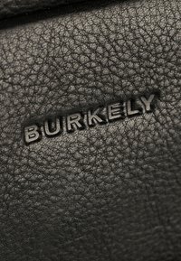Cuir noir texturé avec un grain subtil. Présente le logo "BURKELY" en relief dans une police élégante et moderne. Détail en gros plan.