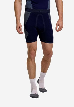 Mann trägt marineblaue, enganliegende Shorts und passendes Oberteil mit weißen, bis zur Mitte der Waden reichenden Socken mit grauen Zehen und Fersen, steht auf hellem Hintergrund.