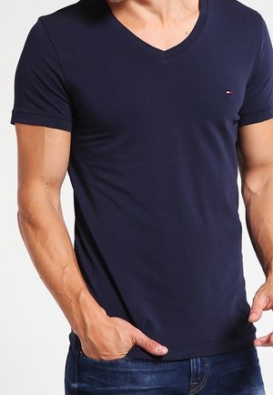 Homme portant un t-shirt bleu marine à manches courtes et col en V avec un petit logo sur la poitrine, assorti à un jean bleu, mains dans les poches.