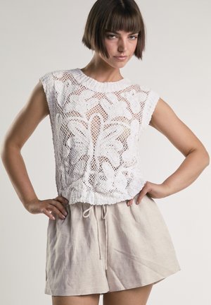 Kvinde med kort brunt hår iført hvid ærmeløs blonder-top med blomsterprint og beige shorts med snøre, poserer med hænderne på hoften.