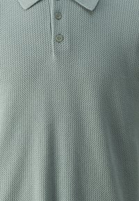 Polo ajusté en tissu maille doux et texturé, de couleur vert clair, avec un col et trois boutons légers le long de la patte de boutonnage.