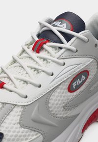 Λευκό αθλητικό παπούτσι FILA με λευκά κορδόνια, κόκκινες λεπτομέρειες, πλευρές από διάτρητο δίχτυ και μπλε σκούρο σήμα με το λογότυπο στη γλώσσα και στην πλευρά.
