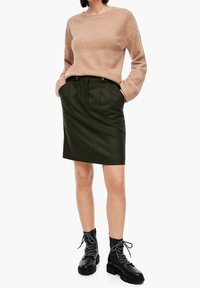 Pull en tricot beige associé à une jupe crayon vert foncé, avec des plis avant, des poches latérales et des bottes de combat noires à lacets.