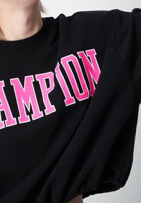 Svart kort sweatshirt med ribbat ok och elastiska ärmar, med djärv, rosa "CHAMPION"-text över bröstet.
