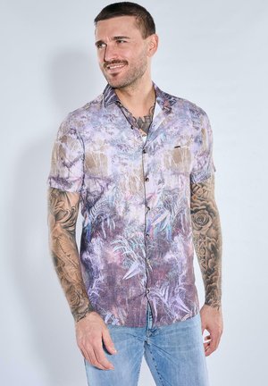 Mann mit Tattoos, der ein kurzärmeliges, knöpfbares Hemd mit lila botanischem Muster und hellblauen Jeans trägt, schaut zur Seite und lächelt.