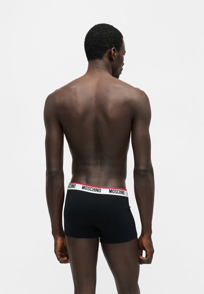 Moški, viden od zadaj, nosi črne boksarice z belim pasom, na katerem je logo Moschino v črni barvi in rdeči poudarki.
