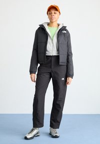 Giacca a vento nera con cappuccio, felpa grigia conzip sotto, pantaloni neri e scarpe da trekking argentate. Un cappello arancione completa l'outfit.