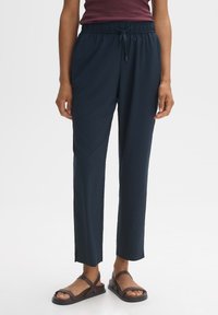 Marineblauwe lichtgewicht broek met een taps toelopende snit, een elastische tailleband en koorddetails. Gecombineerd met bruine sandalen op blote voeten.