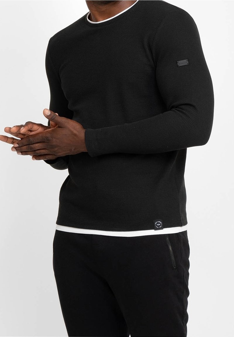 Homme portant un haut noir moulant à manches longues en tissu gaufré superposé à une chemise blanche, assorti d'un pantalon noir avec une poche zippée.