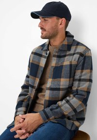 Geruit overhemdjack in marineblauw, grijs en beige. Flanellen materiaal met een knoopsluiting aan de voorkant, kraag en een borstzak. Draagt over een bruine trui.