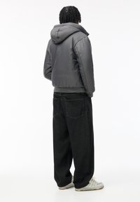 Veste matelassée gris foncé avec capuche ; présente une texture lisse et un design légèrement gonflé. Portée avec un jean noir ample et des baskets blanches.