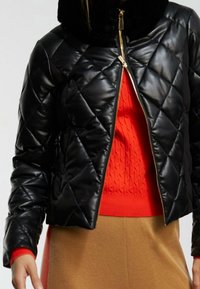 Svart quiltad jacka med glansig finish och en mjuk svart krage, med en guldfärgad dragkedja och en orange texturerad tröja under.