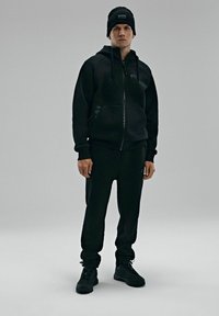 Czarna bluzka z kapturem na zamek i kieszeniami, w komplecie z dopasowanymi czarnymi dresami. Uzupełniona czarną czapką beanie i czarnymi sneakersami.