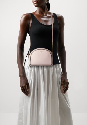 Kvinde i sort ærmeløs top og hvid plisseret nederdel med en lyserød Kate Spade crossbody-taske med guldkæde.