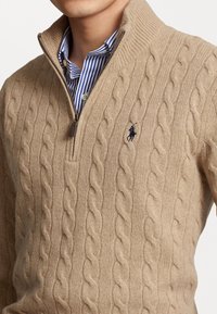 Pull zip en maille torsadée beige porté sur une chemise à rayures bleues et blanches, avec un petit logo sombre brodé sur la poitrine.