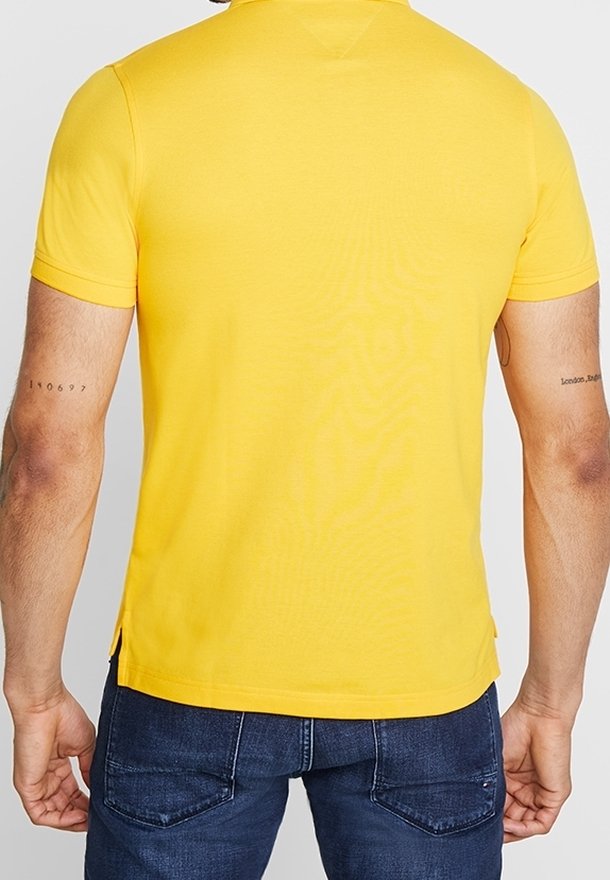 Żółta koszulka polo z krótkim rękawem, kołnierzykiem, rozcięciami po bokach i gładką fakturą, zestawiona z ciemnoniebieskimi dżinsami.