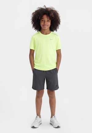 Παιδί με κατσαρά μαλλιά όρθιο, φοράει φούξια-κίτρινη μπλούζα Nike, σκούρα γκρι σορτς και λευκά αθλητικά παπούτσια, με τα χέρια στις τσέπες.