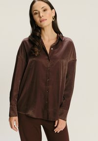 Camisa de satén marrón, de botón y con un ajuste holgado, de manga larga y textura suave. Presenta un cuello y un brillo sutil.