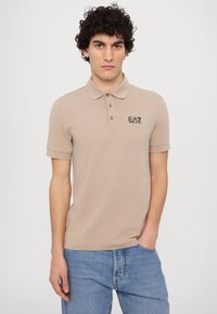 Sandfarget bomulls polo skjorte med krage, tre knapper og svart EA7-logo. Kombinert med blå denimjeans. Glatt tekstur, tettsittende design.